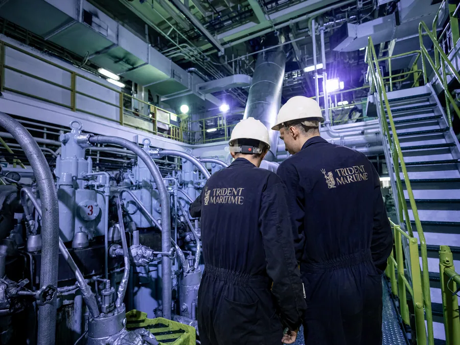Two Trident Maritime superintendents in branded coveralls and hard hats inspecting the main engine in a vessel's engine room.